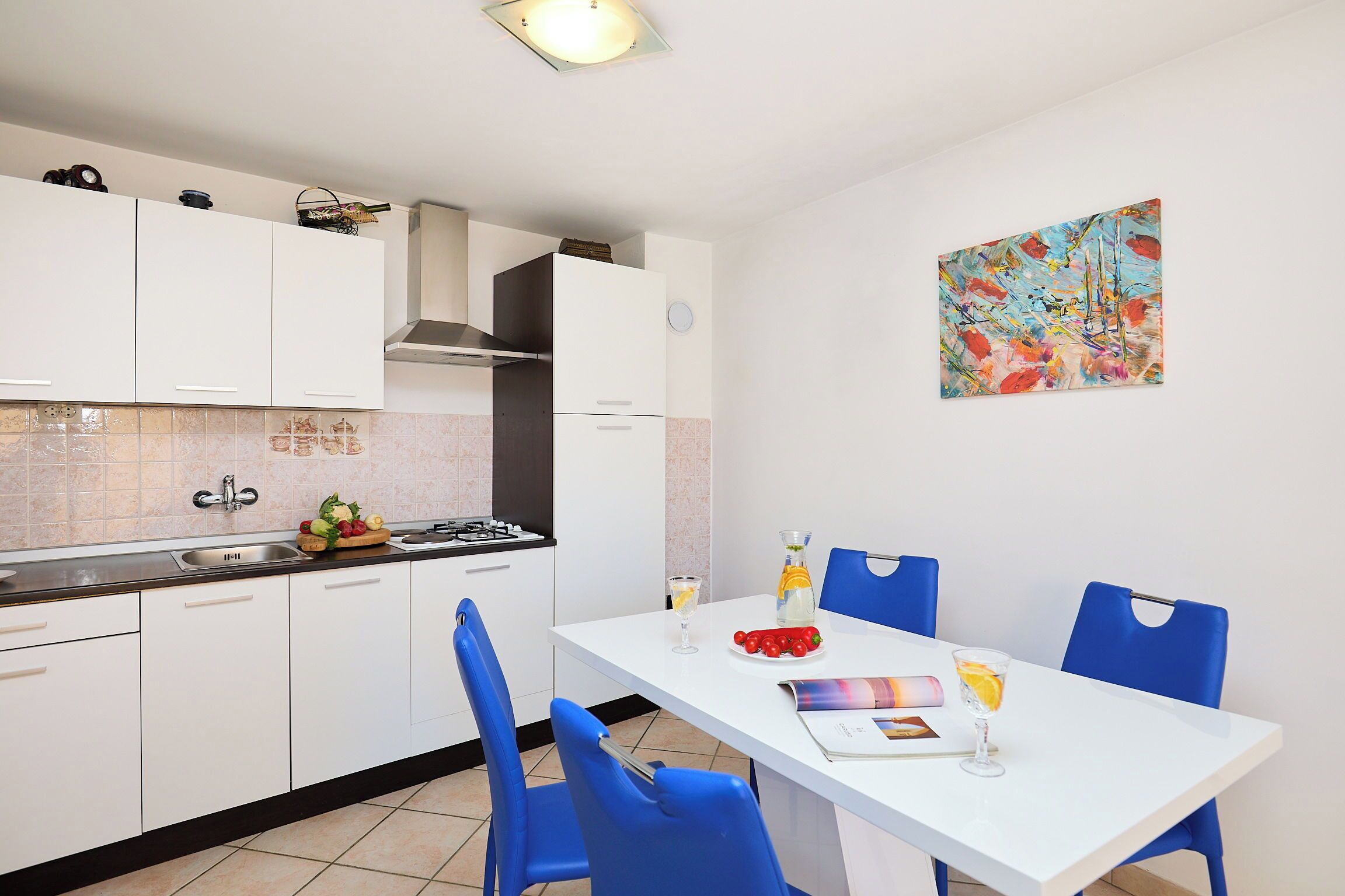 Una cocina moderna con armarios blancos, una mesa de comedor con sillas azules y un cuadro abstracto colorido en la pared.