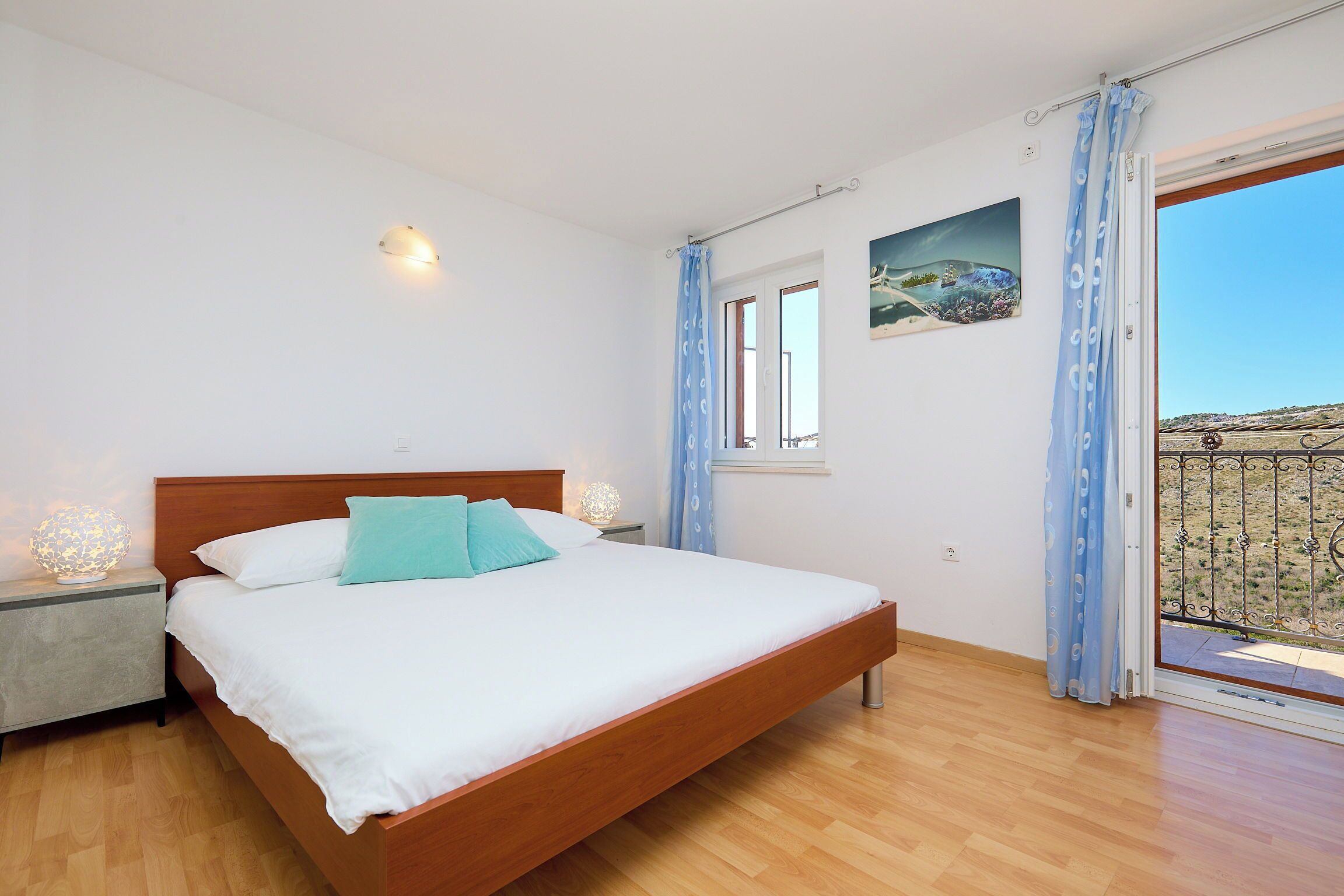 Un dormitorio con una cama de madera, una mesita de noche, una lámpara de pared, un ventanal con cortinas y un balcón con barandilla.