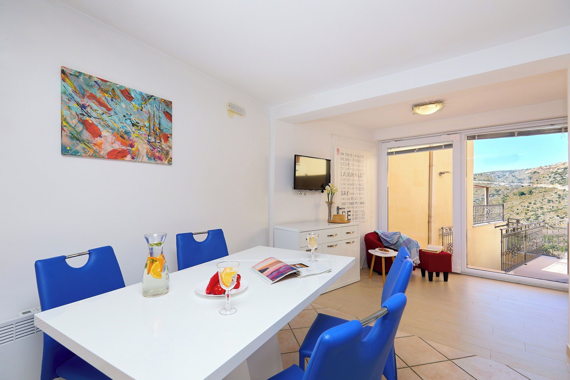 A dining area with a white table, blue chairs, a painting on the wall, and a view through a glass door.