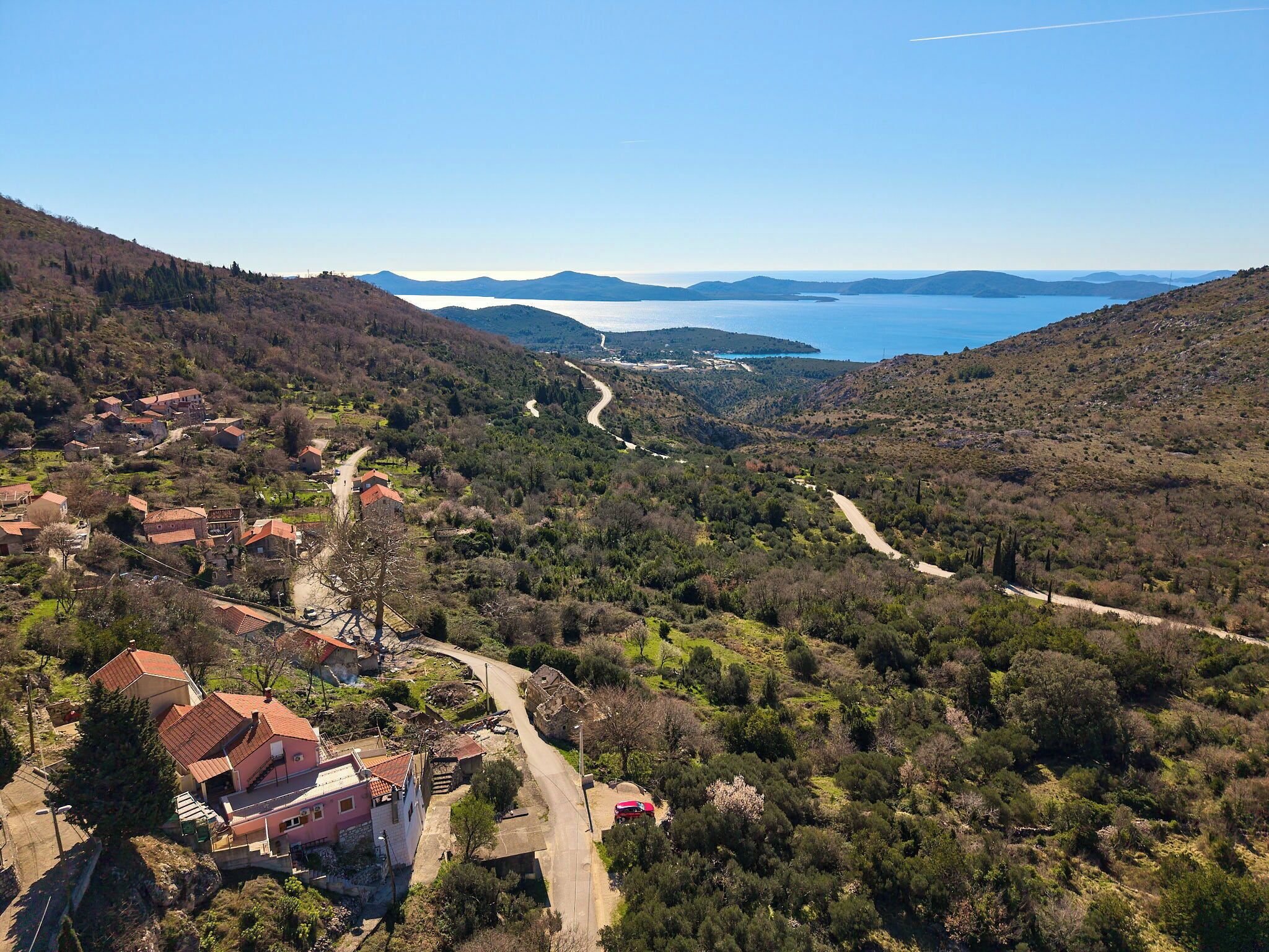 A coastal village with traditional houses, a winding road, and a view of the sea and distant mountains.