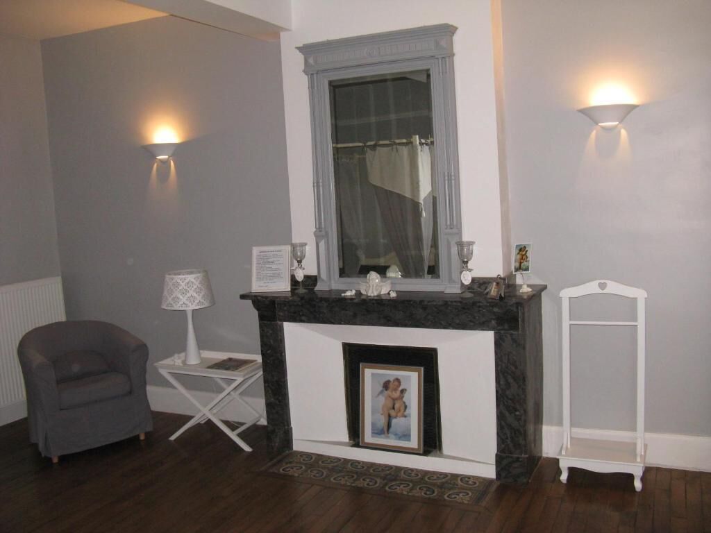 A room with a fireplace, a grey armchair, a small table with a lamp, and a white shelf.