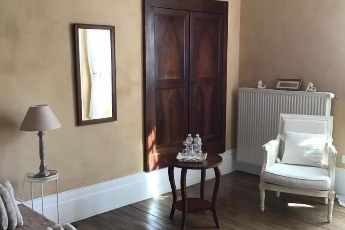 A bedroom with a bed, a chair, a table, a radiator, and a large wooden door.