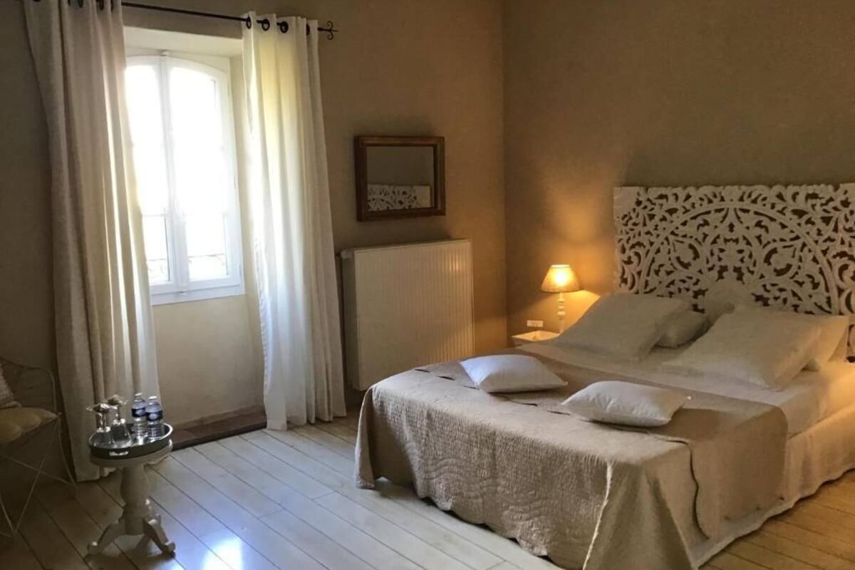 A bedroom with a bed, a bedside table with a lamp, a window with curtains, and a radiator.
