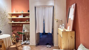 Une petite pièce chaleureuse avec un plancher en bois, un lit avec des oreillers, une commode en bois, une petite table avec une chaise et une étagère ornée d’objets décoratifs.