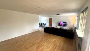 Amplia sala de estar con piso de madera, un sofá negro, un televisor de pantalla plana y un comedor al fondo.