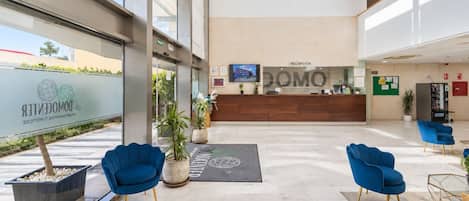 A modern hotel lobby with a reception desk, blue chairs, potted plants, and a large window.