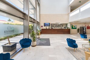 A modern hotel lobby with a reception desk, blue chairs, potted plants, and a large window. - GuestReady - Domocenter studio with garage (Bormujos)