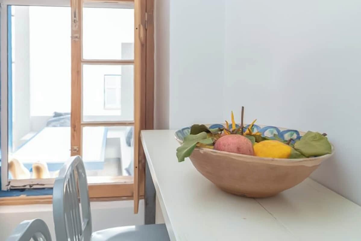 Een kamer met een raam met houten kozijn, een witte tafel en een schaal met fruit.