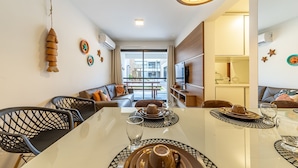 A dining area with a table set for two, a sofa, and a view of the outside. - APTO Próximo à Praia | Barra de São Miguel | San Martin | Destino BSM 106A (Barra de São Miguel)