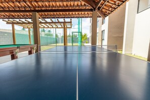 A ping pong table under a wooden pergola with a view of a fenced area and a building in the background. - APTO Próximo à Praia | Barra de São Miguel | San Martin | Destino BSM 106A (Barra de São Miguel)