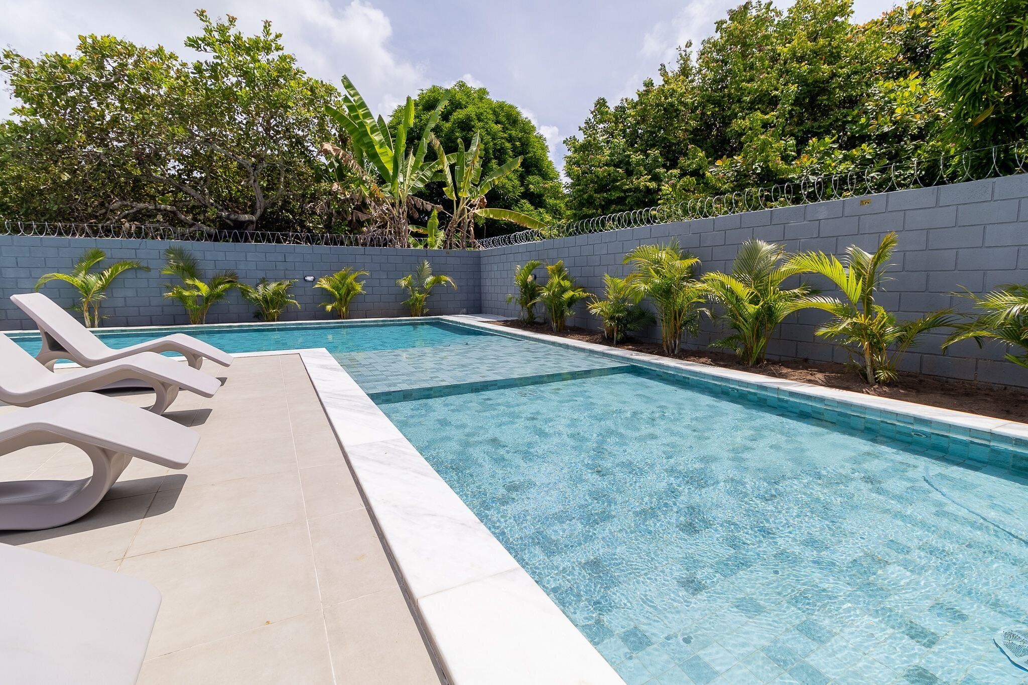 A rectangular swimming pool with clear blue water, surrounded by a low grey wall and a barbed wire fence. There are several lounge chairs along the poolside.