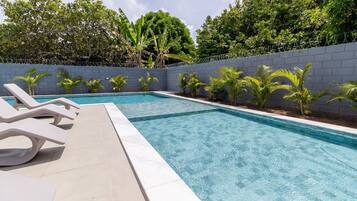 A rectangular swimming pool with clear blue water, surrounded by a low grey wall and a barbed wire fence. There are several lounge chairs along the poolside.