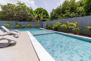 Et rektangulært swimmingpool med klart, blåt vand, omgivet af en lav, grå mur og et pigtrådshegn. Der er flere liggestole langs poolkanten.