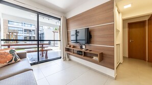 A modern living room with a large TV, wooden shelving, a sofa, and a sliding glass door leading to an outdoor area. - APTO Próximo à Praia | Barra de São Miguel | San Martin | Destino BSM 106A (Barra de São Miguel)
