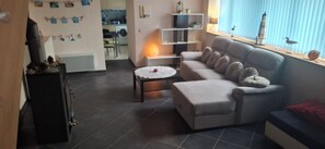 A living room with a grey sofa, a round coffee table, and a small side table. There is a window with blinds and a decorative bird on the sill. - House by the sea (Trévou-Tréguignec)