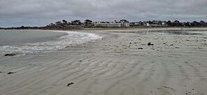 A sandy beach with gentle waves, a distant shoreline with buildings, and a cloudy sky. - House by the Sea (Trévou-Tréguignec)