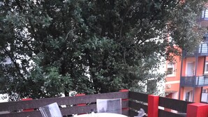 Ein runder Tisch mit vier klaren Plastikstühlen auf einer Holzterrasse, umgeben von einem Holenzaun mit roten Akzenten.