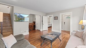 A spacious living area with a wooden floor, a large flat-screen TV, a sofa, a coffee table, and a dining area with chairs. - Spacious Downtown Retreat w/ Private Yard & Mountain Views (Colorado Springs)