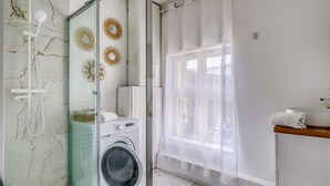Ein modernes Badezimmer mit Duschkabine aus Glas, einer Waschmaschine und einem Fenster mit durchsichtigen Vorhängen.