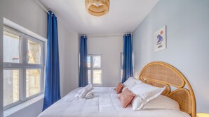 Ein ordentlich bezogenes Bett mit weißen und rosafarbenen Kissen, einem Korbmöbel-Kopfstück und einem Fenster mit blauen Vorhängen.