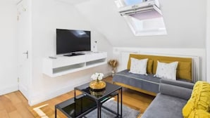 A modern living room with a flat-screen TV on a white console, a sofa with yellow cushions, and a small table with a decorative item. - 4BR London Home Piccadilly Line Sleeps 9 (London)