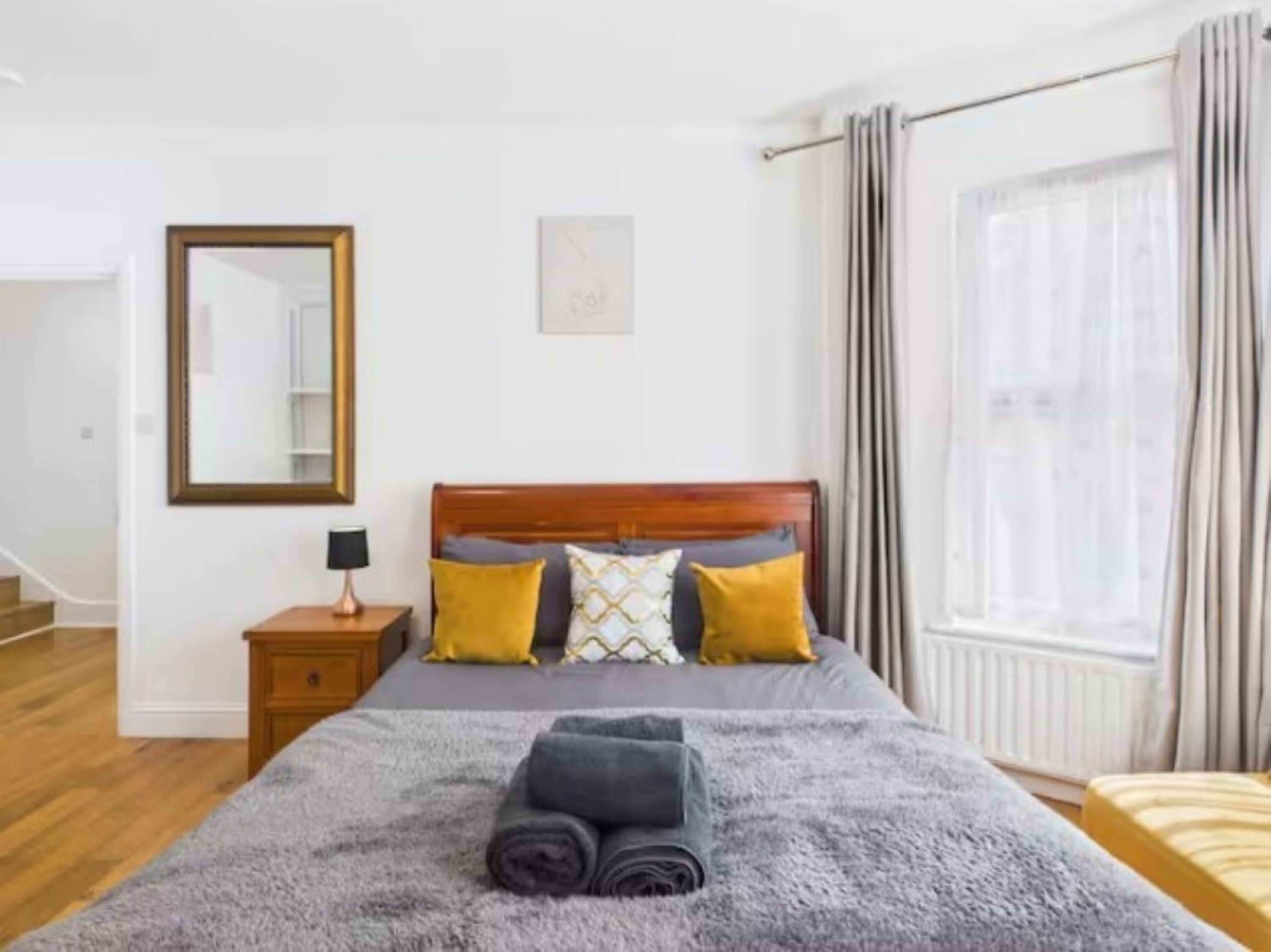 Un dormitorio con una cama de madera, una mesita de noche con lámpara, un espejo y una ventana con cortinas.