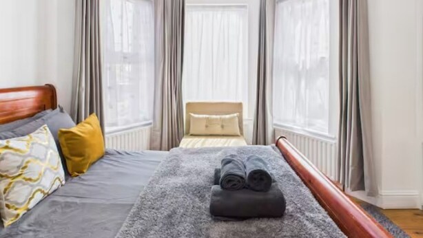 A hotel room with a bed, a chair, and a view of the city through large windows. - 4BR London Home Piccadilly Line Sleeps 9 (London)