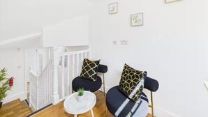 A modern living room with a staircase, a black sofa with patterned cushions, a small white table with a plant, and framed artwork on the wall. - 4BR London Home Piccadilly Line Sleeps 9 (London)
