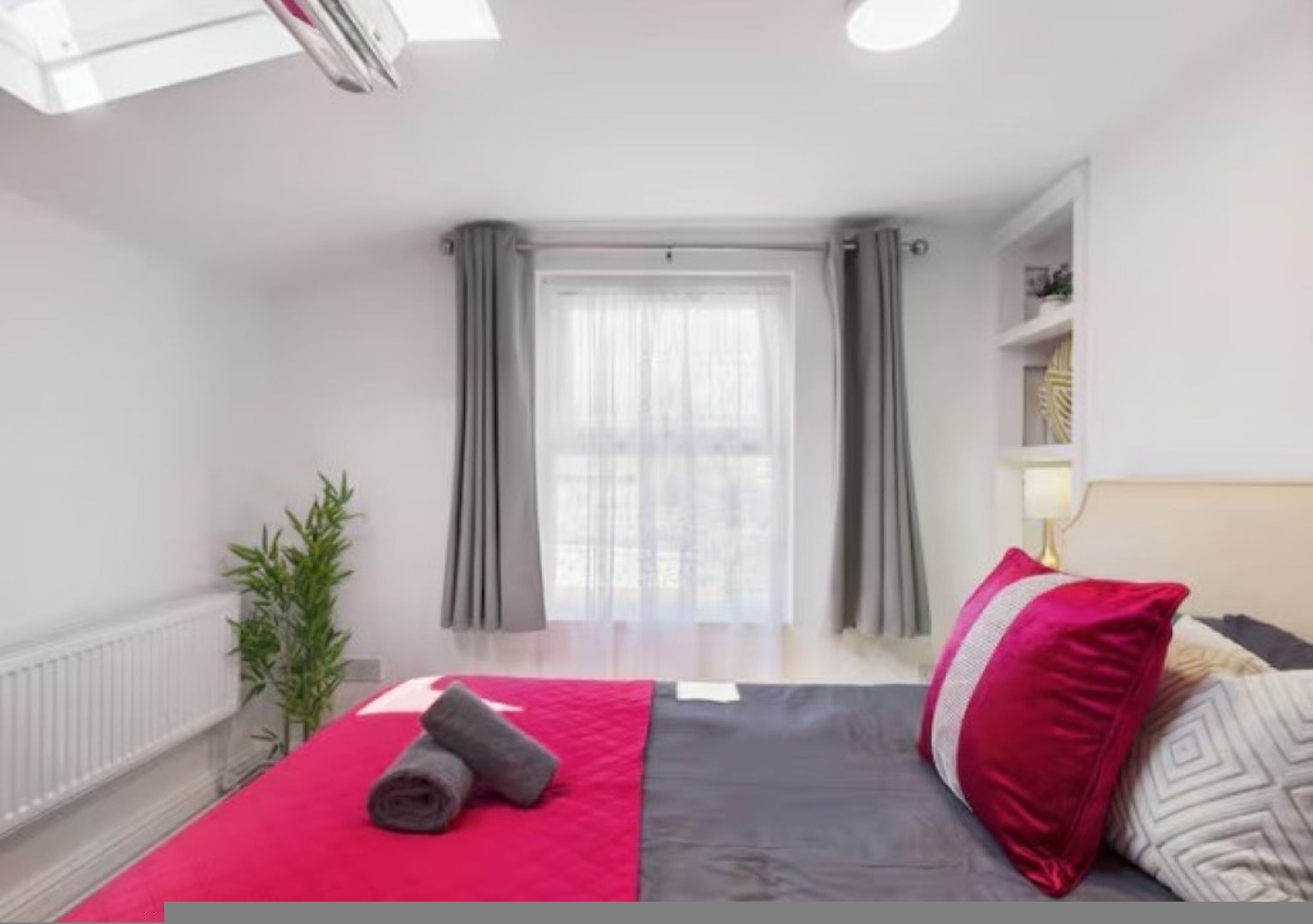 A modern hotel room with a bed, a window with curtains, a bedside table with a lamp, and a small plant.