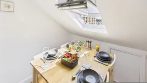 A dining area with a table set for two, a basket of fruit, and a view of the city through a skylight. - 4BR London Home Piccadilly Line Sleeps 9 (London)