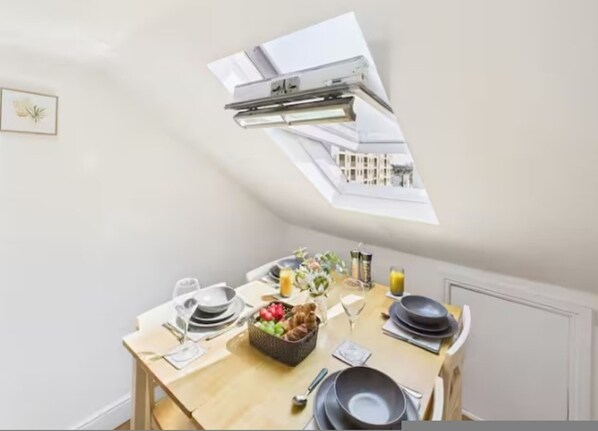 Un comedor con una mesa dispuesta para dos, una cesta de fruta y vistas de la ciudad a través de un tragaluz.