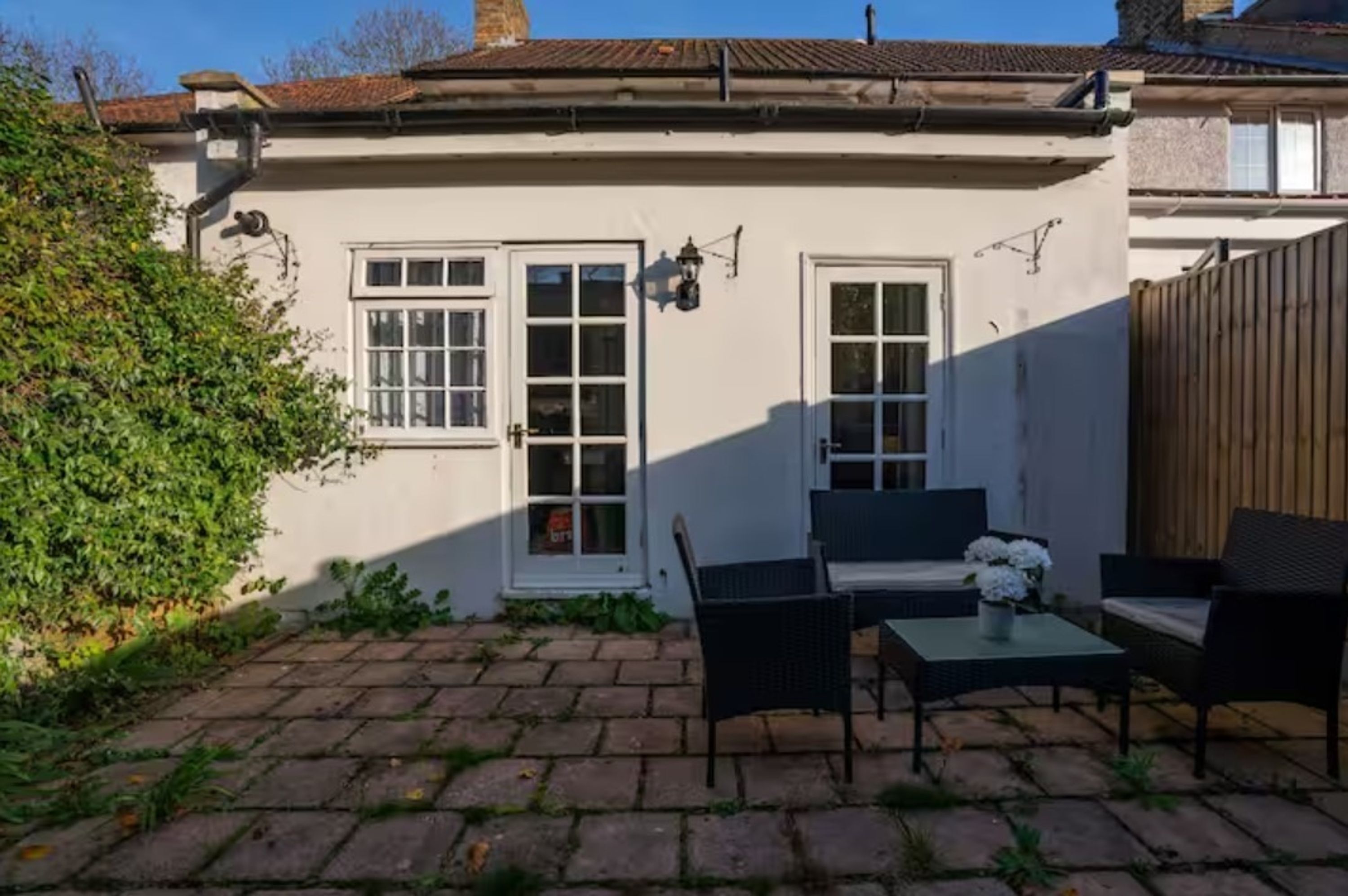 A white building with two windows and a door, a paved patio with outdoor furniture, and a wooden fence.