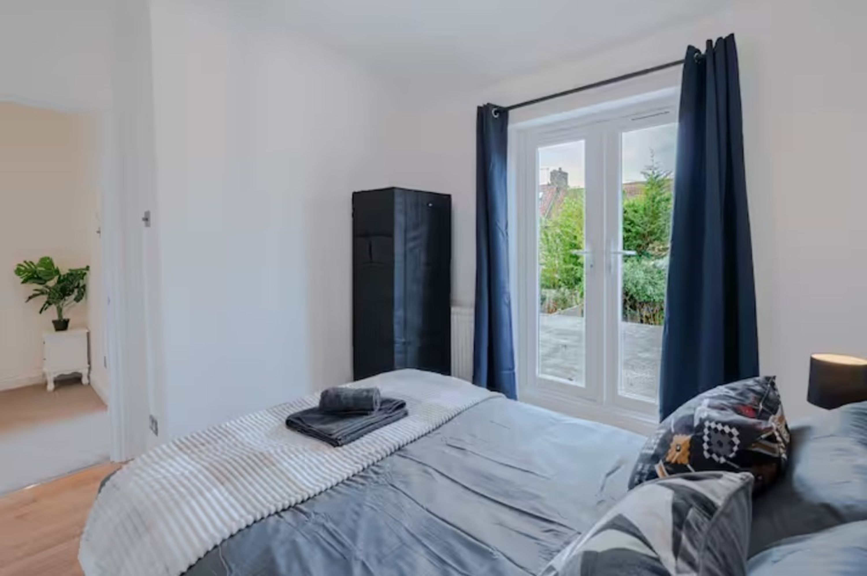 A bedroom with a bed, a window with curtains, a potted plant, and a black cabinet.