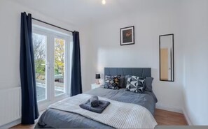 A bedroom with a bed, a nightstand, a lamp, a framed picture, and a window with curtains. - 3BR Putney Home London Sleeps 8 Winter Deal (London)