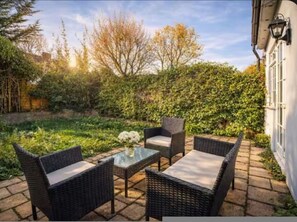 An outdoor patio with wicker furniture and a glass table, surrounded by lush greenery and a white building wall. - 3BR Putney Home London Sleeps 8 Winter Deal (London)