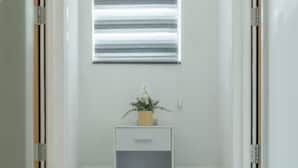 Un couloir avec une fenêtre dotée de stores horizontaux, une petite table de chevet blanche ornée d’une plante, et une porte sur la droite.