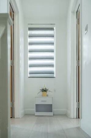 A hallway with a window with horizontal blinds, a small white bedside table with a plant, and a door on the right. - Family-friendly 3BR Home London N17 Sleeps 8 (London)