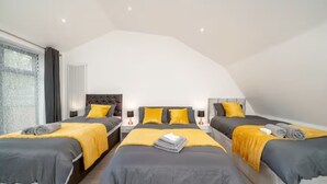 Une chambre moderne avec trois lits, chacun recouvert d’une couvre-lit grise et jaune. On y trouve des tables de chevet avec des lampes, une fenêtre avec des rideaux et un radiateur.