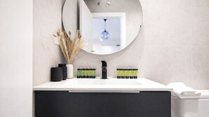 Un baño moderno con un lavamanos blanco, un mueble negro y un espejo redondo. Hay plantas decorativas y una toalla en la encimera.