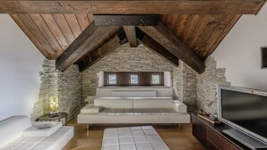 A loft-style room with exposed wooden beams, a stone wall, a white sofa, a television, and a small table with a lamp.