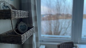 Una habitación con estantes de mimbre que sostienen objetos decorativos, vista de un paisaje nevado a través de una ventana, y un sillón de mimbre con cojín.