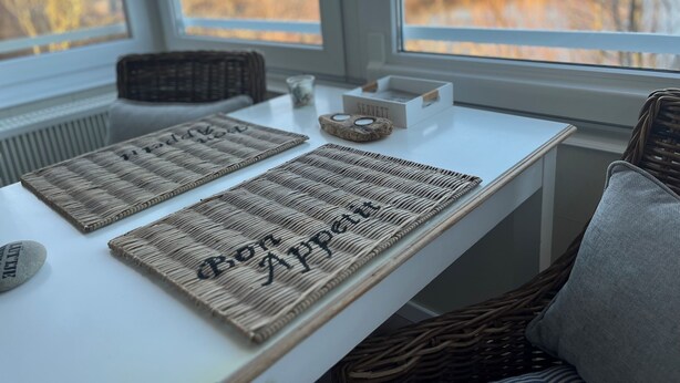 Una mesa con dos individuales tejidos y una caja pequeña con el rótulo "SERVETT" sobre una mesa blanca en una habitación con ventanas amplias.