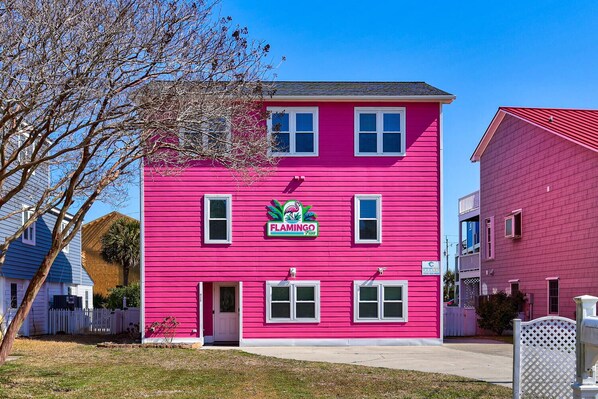 Ein zweistöckiges Gebäude mit einer leuchtend rosa Außenfassade, einem Schild mit der Aufschrift „FLAMINGO FLU“ und einem weißen Ziergartenzaun vor der Fassade.