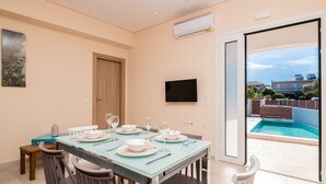 A dining area with a table set for four, a TV mounted on the wall, and a view of a swimming pool through an open door. - Villa Ocean Pool in Planos (Zakynthos)