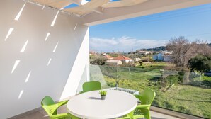 A balcony with a white table and green chairs, overlooking a grassy area with houses and a body of water. - Villa Ocean Pool in Planos (Zakynthos)