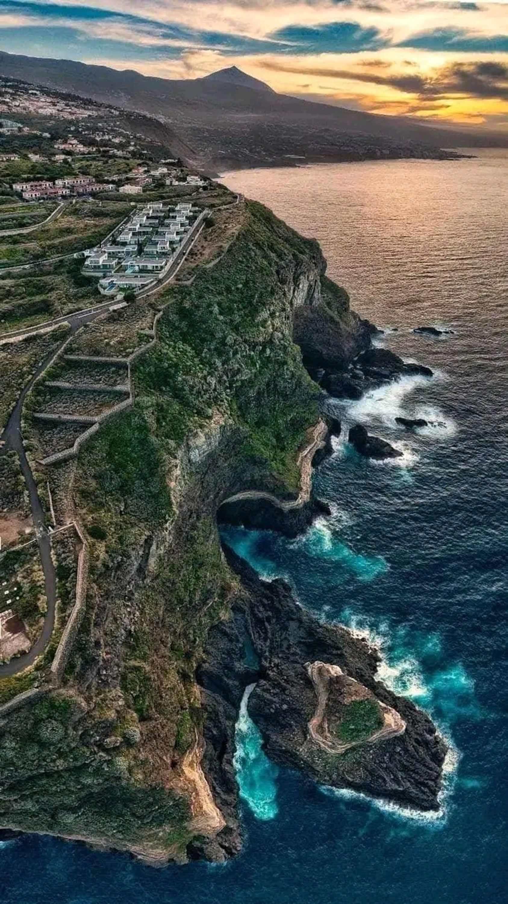 An aerial view of a coastal area with a terraced structure, a winding road, and a coastline with rocky outcrops and turquoise waters.