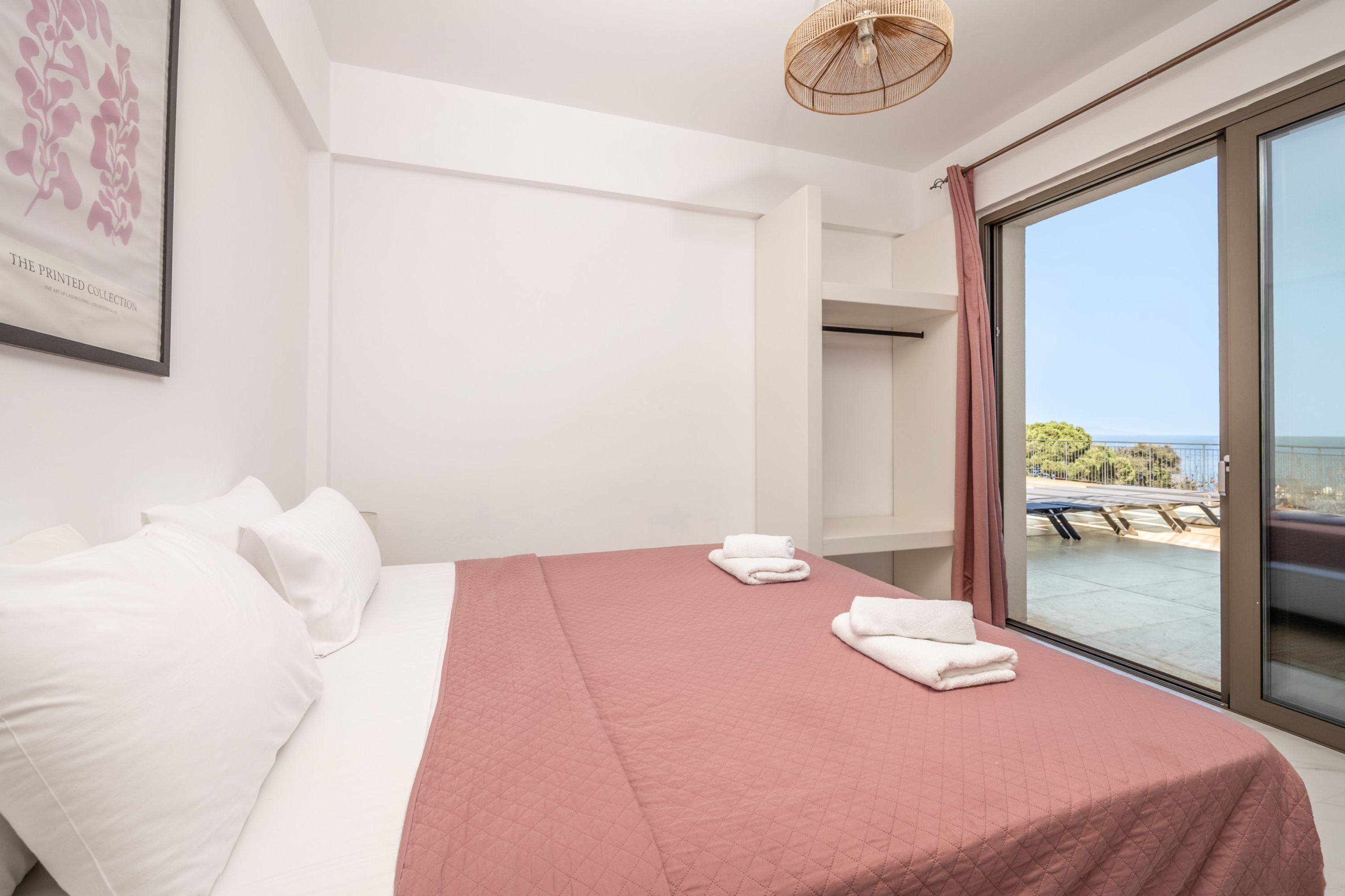 A hotel room with a bed, white pillows, a pink bedspread, and a large window with a view of the sea.
