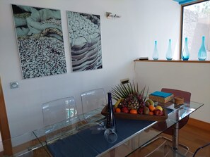 A modern dining area with a glass table, chairs, and a fruit basket. There are two wall art pieces above the table. - Your Island Villas Mamma Mia 2 (Yaiza)