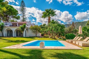 A swimming pool with a clear blue water, surrounded by a well-maintained lawn, palm trees, and a building with a balcony. - Villa Hacienda Zante (Zakynthos)