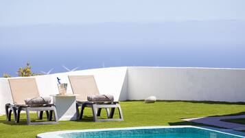 A pool area with two lounge chairs, a small table with a drink, and a view of the ocean.